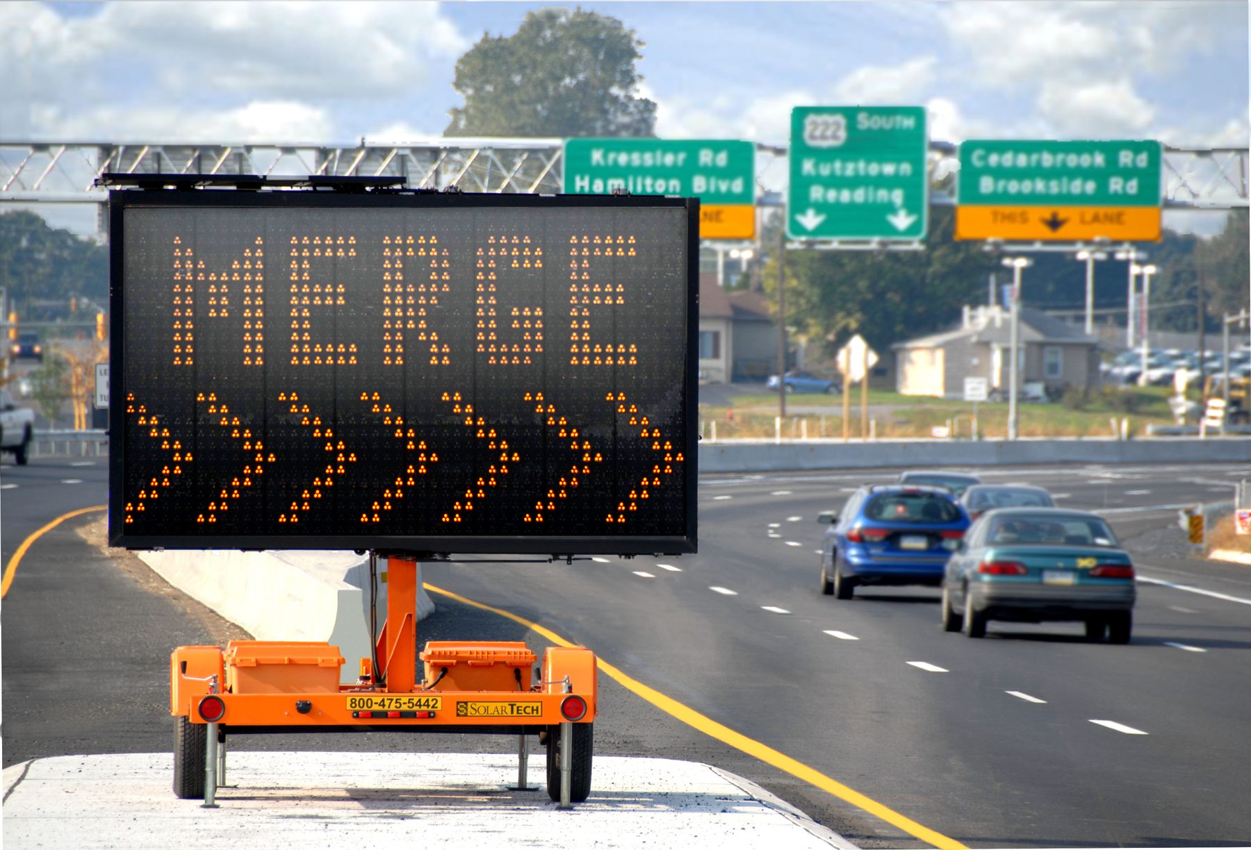 A SolarTech message board on a highway shoulder reading Merge, with traffic passing beside it